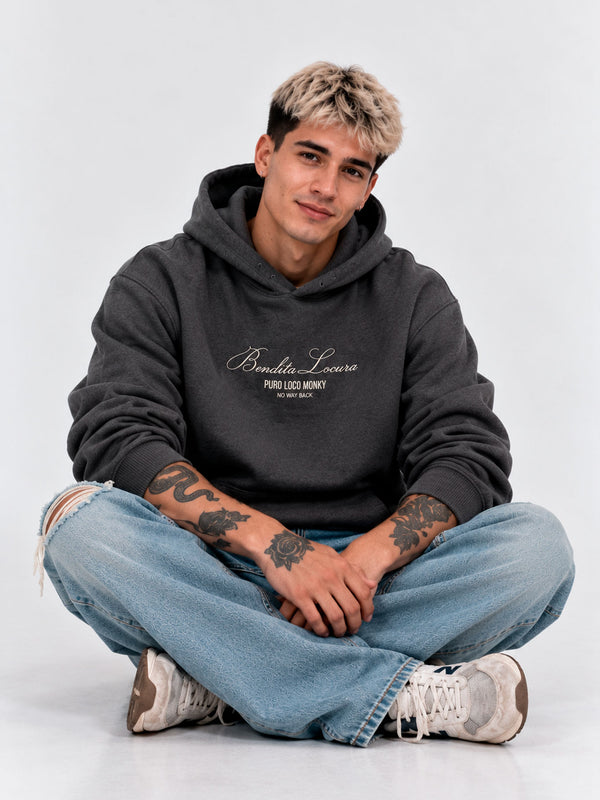 Sudadera unisex gris oscuro con bordado 'Bendita Locura' y logo 'Puro Loco Monky', modelo sentado con jeans claros.