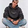 Sudadera unisex gris oscuro con bordado 'Bendita Locura' y logo 'Puro Loco Monky', modelo sentado con jeans claros.