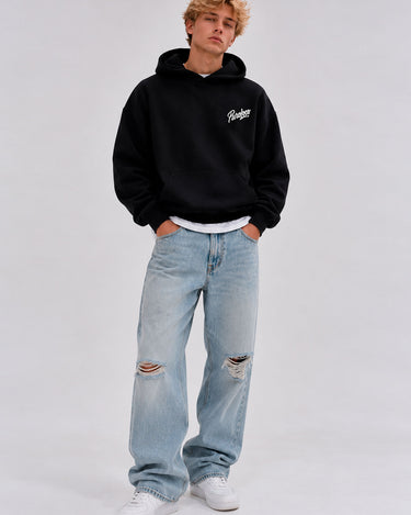Sudadera oversize negra con capucha y logo en el pecho, combinada con jeans claros desgastados y zapatillas blancas.