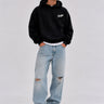 Sudadera oversize negra con capucha y logo en el pecho, combinada con jeans claros desgastados y zapatillas blancas.