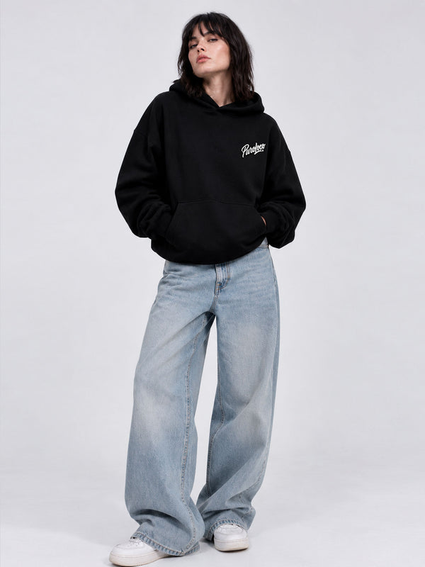 Sudadera oversize negra con capucha, logo blanco en el pecho, combinada con jeans claros y zapatillas blancas.