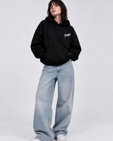 Sudadera oversize negra con capucha, logo blanco en el pecho, combinada con jeans claros y zapatillas blancas.