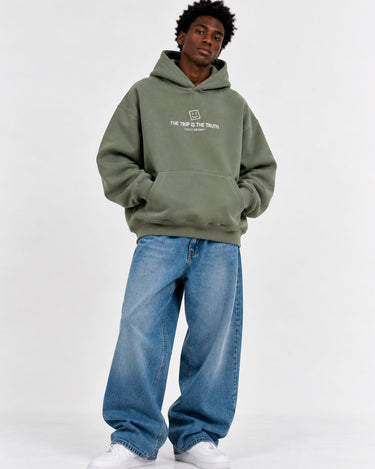 Sudadera boxy unisex en color oliva con capucha y bolsillo frontal, texto en blanco en el pecho, jeans azules.