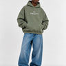 Sudadera boxy unisex en color oliva con capucha y bolsillo frontal, texto en blanco en el pecho, jeans azules.