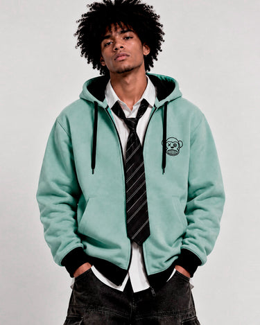 Sudadera con cremallera color verde suave, con capucha y logo de mono en el pecho, combinada con camisa y corbata.