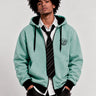 Sudadera con cremallera color verde suave, con capucha y logo de mono en el pecho, combinada con camisa y corbata.