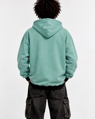 Sudadera con capucha de color verde suave, vista trasera, con pantalones oscuros y cabello rizado.