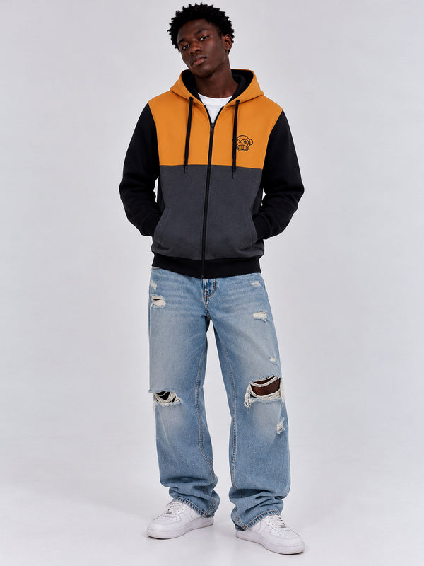 Sudadera con cremallera en mostaza, negro y gris oscuro, con capucha y logo en el pecho, modelo masculino con jeans rotos.