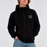 Sudadera negra con cremallera, capucha y logo de mono en el pecho, modelo masculino con jeans claros.