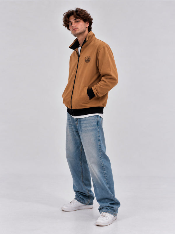 Chaqueta sin capucha color caramelo con logo en el pecho, combinada con jeans claros y zapatillas blancas.
