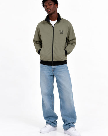 Chaqueta sin capucha color oliva con cierre, logo en el pecho, combinada con jeans claros y zapatillas blancas.