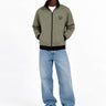Chaqueta sin capucha color oliva con cierre, logo en el pecho, combinada con jeans claros y zapatillas blancas.