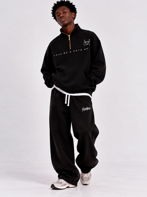 Sudadera oversize negra con cremallera y texto 'LOVE ME & HATE ME', pantalones negros, modelo de pie.