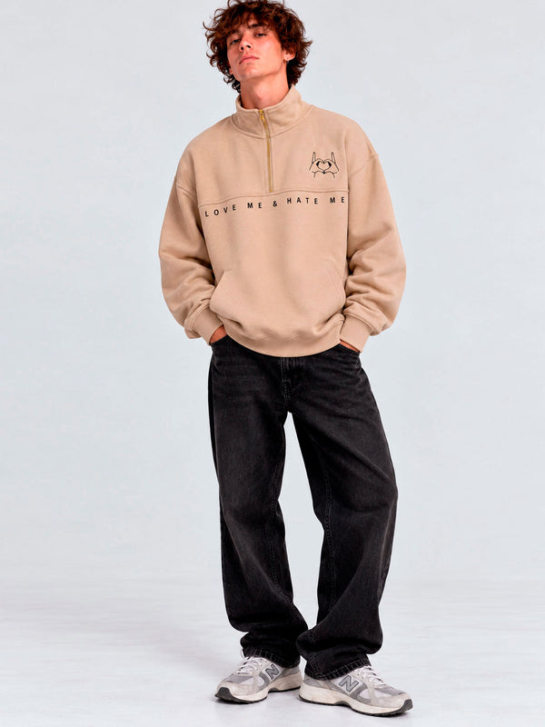 Sudadera beige con cremallera, logo bordado en el pecho, texto 'LOVE ME & HATE ME', pantalones negros.