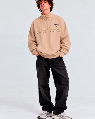 Sudadera beige con cremallera, logo bordado en el pecho, texto 'LOVE ME & HATE ME', pantalones negros.