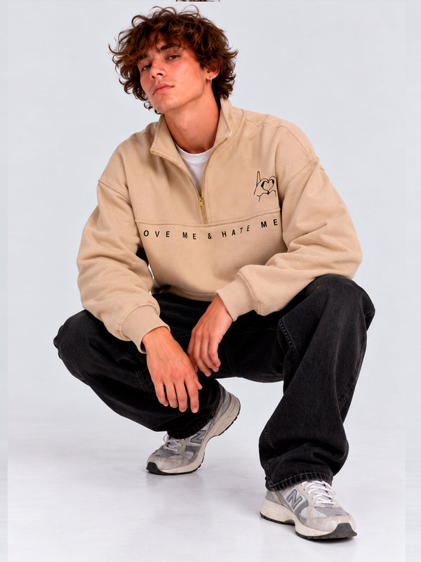 Sudadera oversize beige con logo bordado 'LOVE ME & HATE ME' en el pecho, modelo sentado, jeans negros.