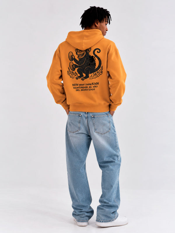 Sudadera unisex mostaza con diseño de mono y texto en la parte posterior, jeans claros y fondo neutro.