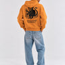 Sudadera unisex mostaza con diseño de mono y texto en la parte posterior, jeans claros y fondo neutro.