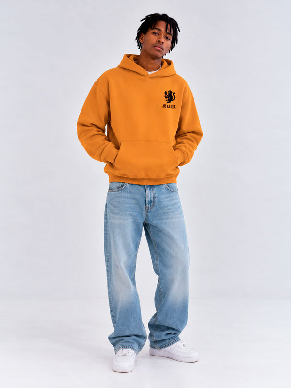 Sudadera unisex color mostaza con capucha y logo negro en el pecho, combinada con jeans claros.