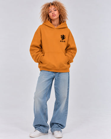 Sudadera unisex color mostaza con capucha, logo negro en el pecho, modelo con pantalones anchos y zapatillas.