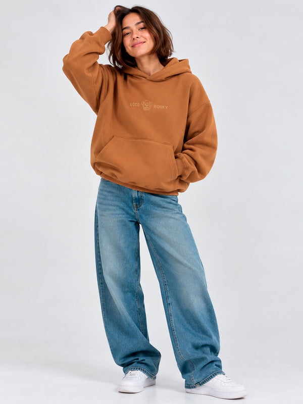 Sudadera oversize marrón con capucha y logo de Loco Monky, combinada con jeans claros y zapatillas blancas.