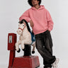 Sudadera rosa con capucha y logo de cara de mono, modelo posando junto a un caballito de madera.