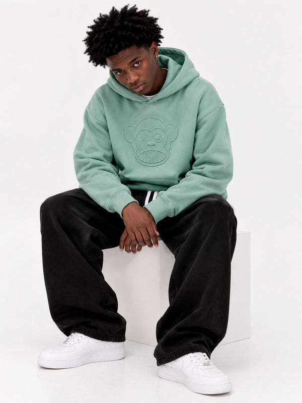 Sudadera unisex verde suave con capucha y diseño de mono en el pecho, pantalones negros y zapatillas blancas.