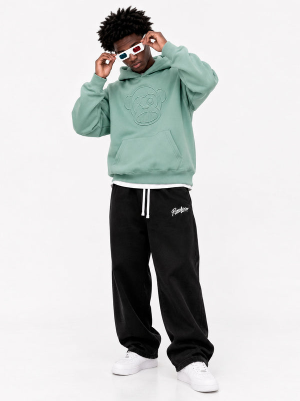 Sudadera verde suave con capucha y diseño de mono en relieve, pantalones negros y gafas de sol coloridas.