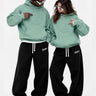 Sudadera verde suave con diseño 3D en el pecho, modelo unisex, combinada con pantalones negros.