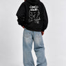 Sudadera negra con capucha, diseño gráfico en la espalda, jeans claros, modelo de pie de espaldas.