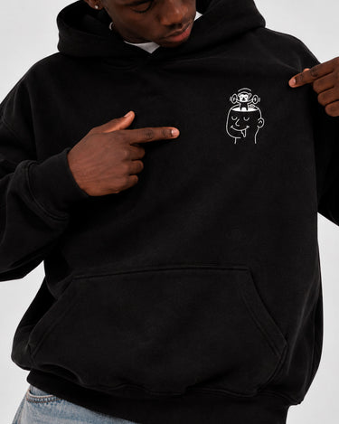Sudadera negra con capucha, diseño gráfico en el pecho, persona señalando el logo.