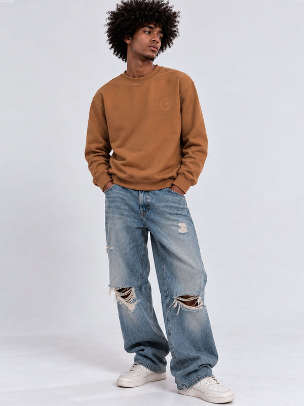 Sudadera color caramelo con logo en el pecho, jeans desgastados y zapatillas blancas, fondo gris claro.
