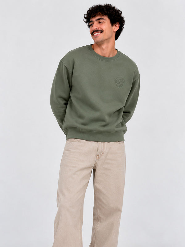Sudadera de color oliva con logo en el pecho, modelo masculino con pantalones claros y fondo neutro.