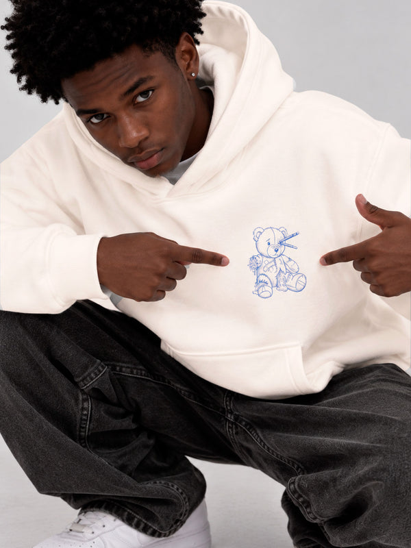 Sudadera blanca unisex con un diseño de oso de peluche azul en el pecho, modelo señalando el diseño.