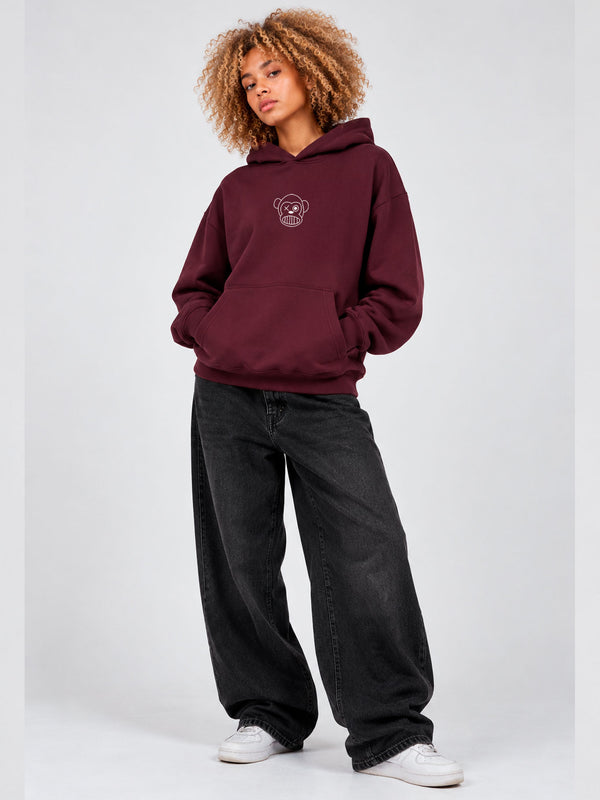Sudadera unisex color burdeos con capucha y logo de mono en el pecho, combinada con pantalones negros amplios.